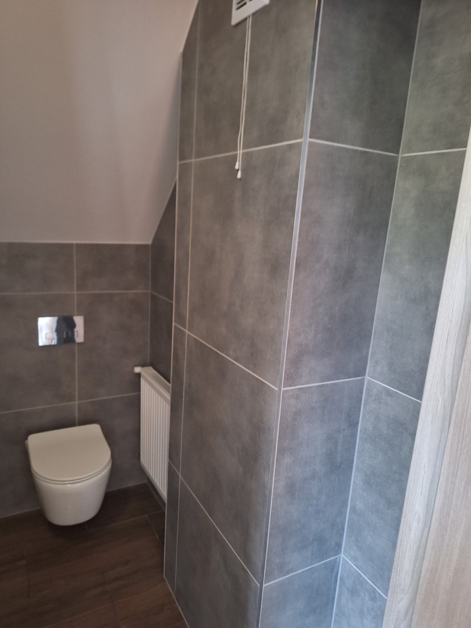 Nowoczesna łazienka z szarymi płytkami, podwieszaną toaletą i grzejnikiem drabinkowym na tle skośnego sufitu.