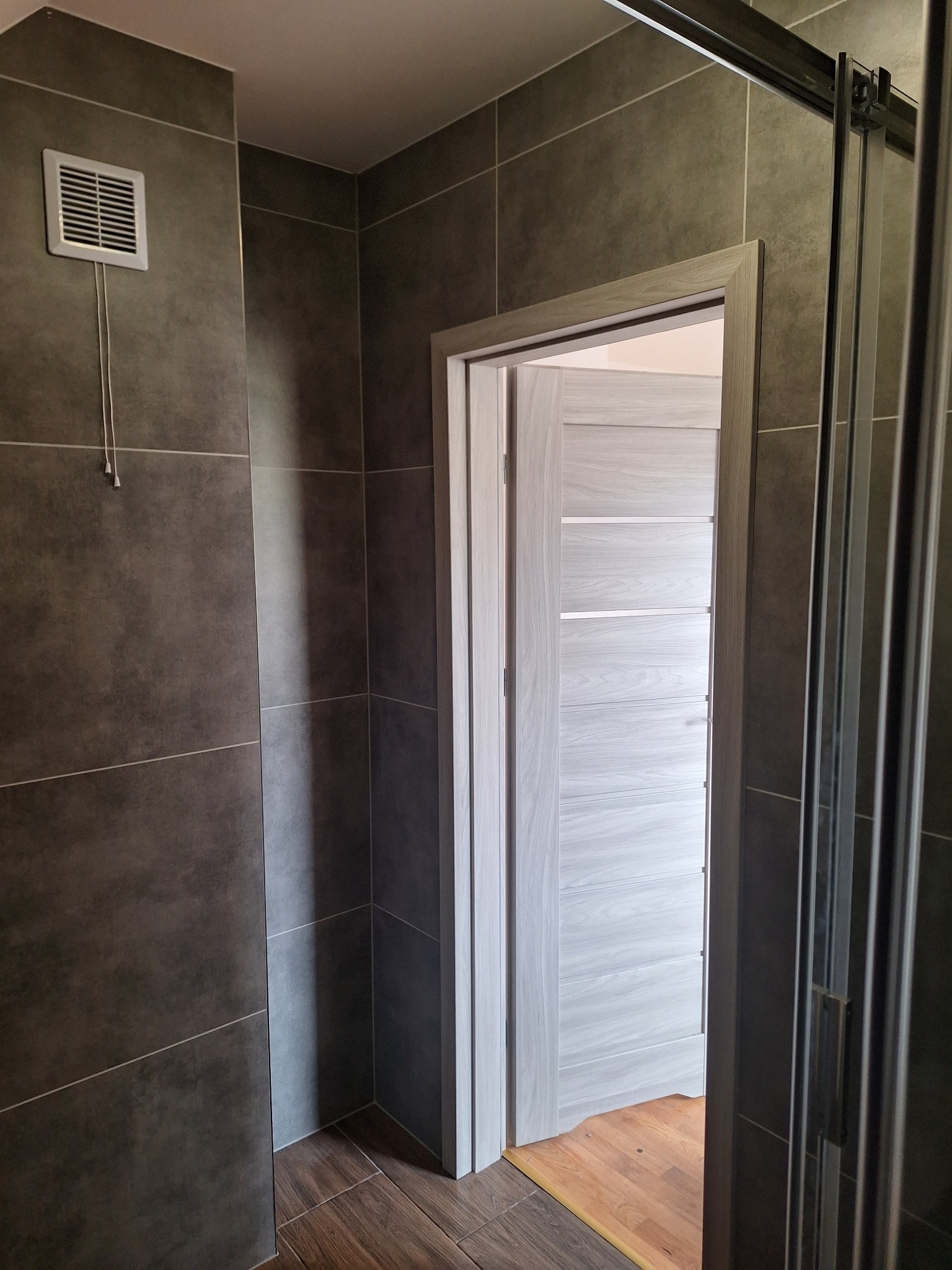 Nowoczesna łazienka z szarymi płytkami, drewnopodobną podłogą i jasnoszarymi drzwiami z poziomymi frezami, widoczny fragment kabiny prysznicowej i kratka wentylacyjna.