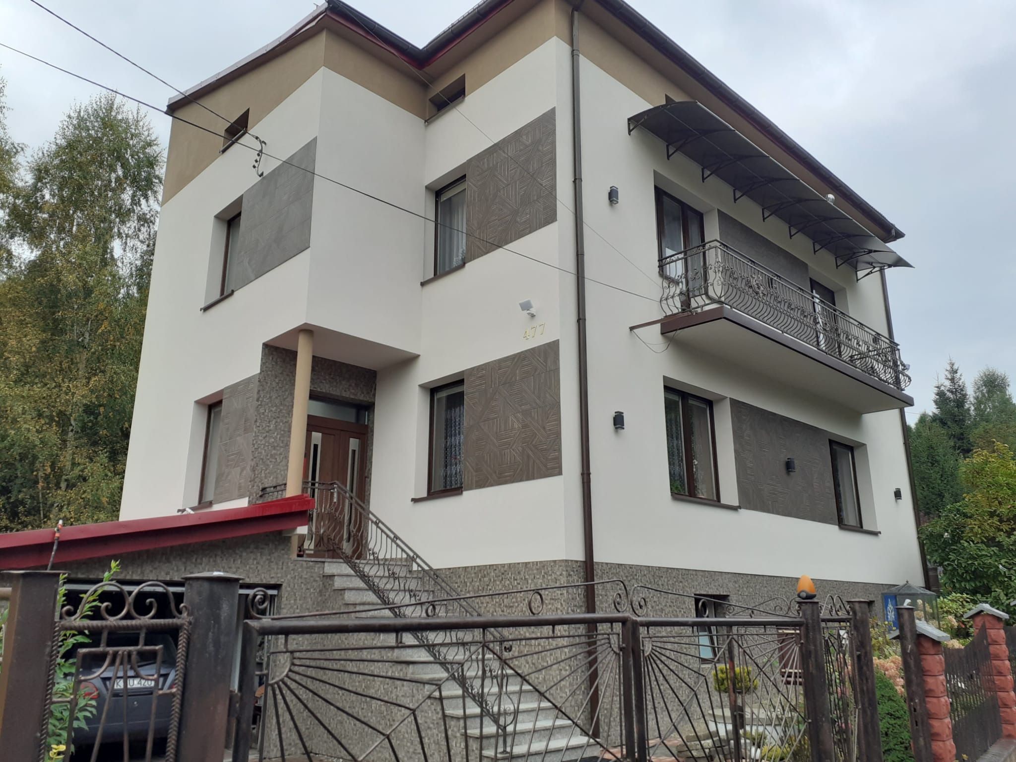 Dwupiętrowy dom z jasną elewacją, dekoracyjnymi panelami i balkonem z kutą balustradą, widziany zza metalowego ogrodzenia.