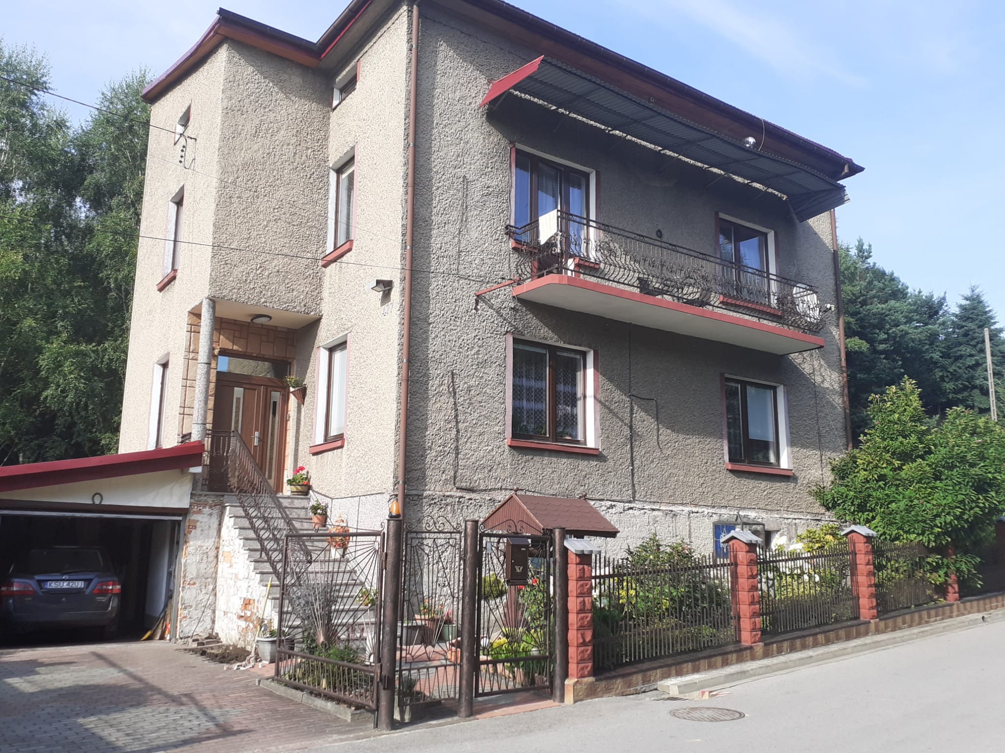 Dwupiętrowy dom z szarą elewacją o fakturze baranka, ciemnoczerwonymi obramowaniami okien i balkonem z metalową balustradą, widok z zewnątrz na garaż z zaparkowanym samochodem.