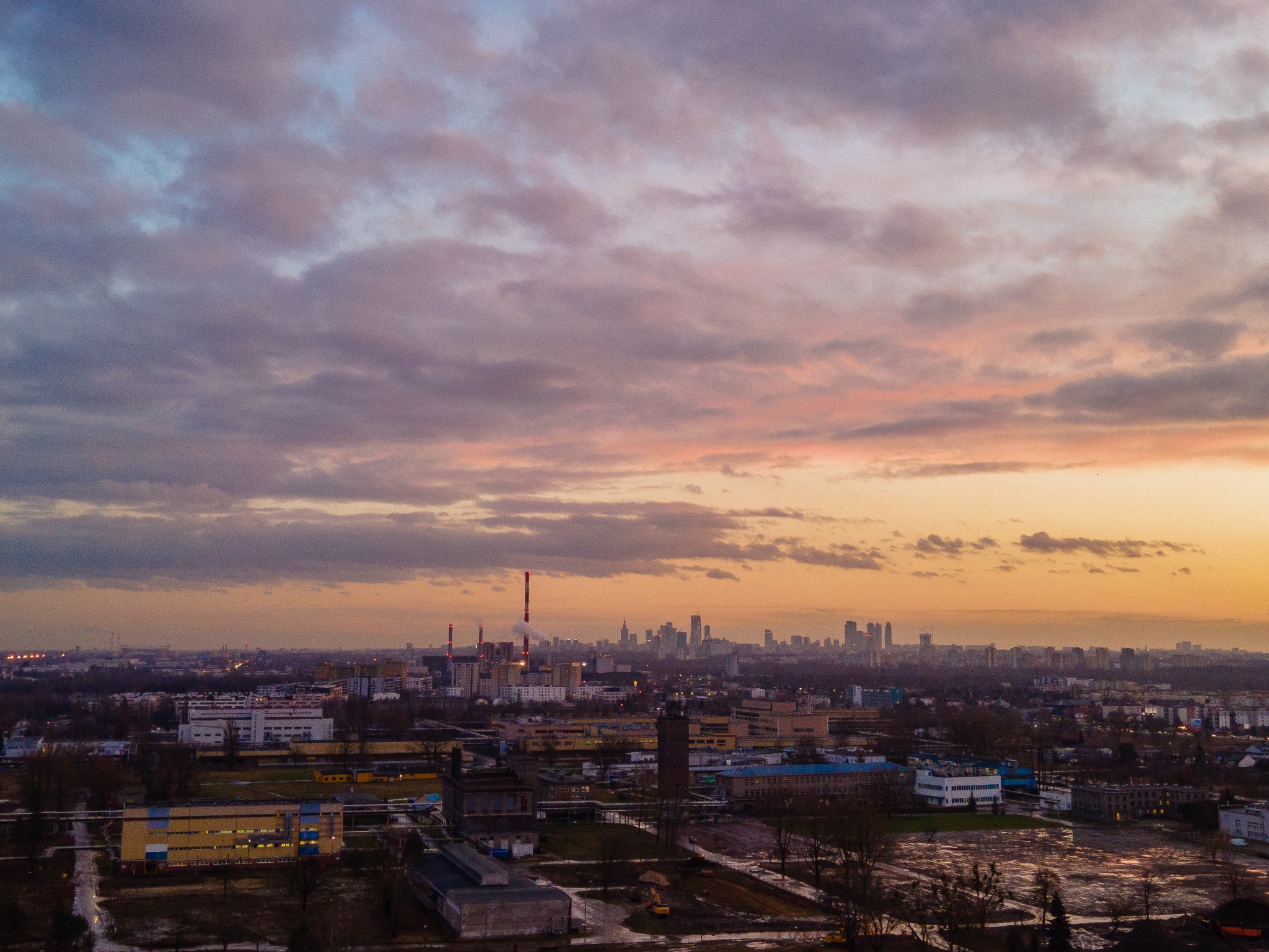Panorama miasta z widokiem na fabrykę i drapacze chmur w oddali, sfotografowana o zmierzchu pod zachmurzonym niebem.
