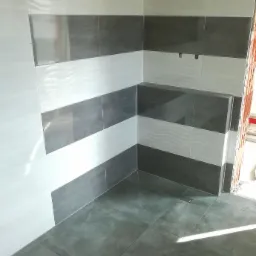 Wykończony narożnik łazienki z ułożonymi płytkami w poziome pasy: szare płytki podłogowe, ściany w biało-szare pasy z delikatnym wzorem fal na białych płytkach, widoczny fragment surowej ściany...