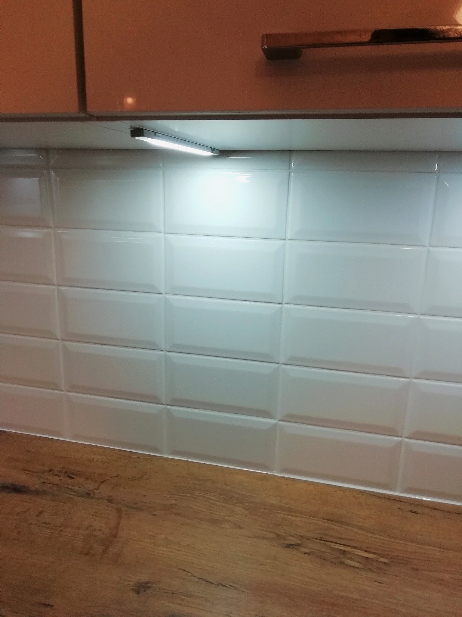 Białe, wypukłe płytki ułożone w rzędach na ścianie nad drewnopodobnym blatem, oświetlone listwą LED pod szafkami wiszącymi.