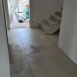 Szare płytki podłogowe ułożone w kwadraty z wąskimi fugami, widoczne schody w surowym stanie i materiały budowlane w tle, zabezpieczone folią drzwi.