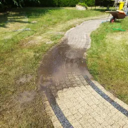 Fragment kostki brukowej w ogrodzie, częściowo umyty, z widocznymi śladami wody i błota, taczka i żółty kosz na śmieci w tle.