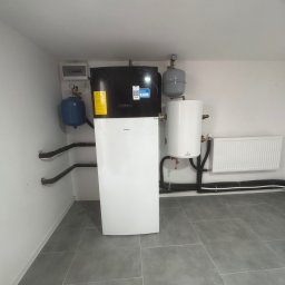 HydroKlim - Nowoczesna instalacja z pompą ciepła Daikin, zbiornikiem buforowym i grzejnikiem panelowym w pomieszczeniu technicznym w Ligocie Małej, z widocznymi czarnymi rurami instalacyjnymi.