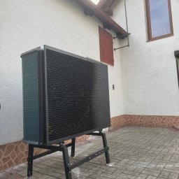 HydroKlim - Czarna pompa ciepła powietrze-woda na stalowym stojaku, zainstalowana przy białej ścianie domu z brązowymi elementami wykończeniowymi, widoczna kostka brukowa.