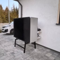 HydroKlim - Zewnętrzna jednostka pompy ciepła powietrze-woda, zainstalowana przy ścianie budynku z widocznymi rurami w izolacji, na tle szarej kostki brukowej i białego samochodu.