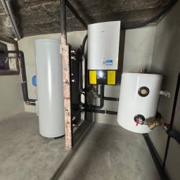 HydroKlim - Instalacja z białym zbiornikiem ciepłej wody, szarym zasobnikiem i pompą ciepła Daikin na tle surowego betonowego sufitu w pomieszczeniu technicznym.