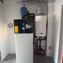 Daikin i Ferroli: zainstalowana pompa ciepła z zbiornikiem buforowym w surowym pomieszczeniu, widoczne rury, manometry, izolowany przewód wentylacyjny pod sufitem, w tle fragment ściany w stanie...