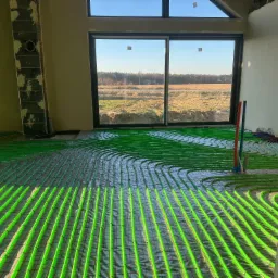 Wnętrze w stanie surowym z widoczną instalacją ogrzewania podłogowego z rur w kolorze jaskrawej zieleni, ułożonych na siatce w nowo budowanym domu z dużym oknem na krajobraz.