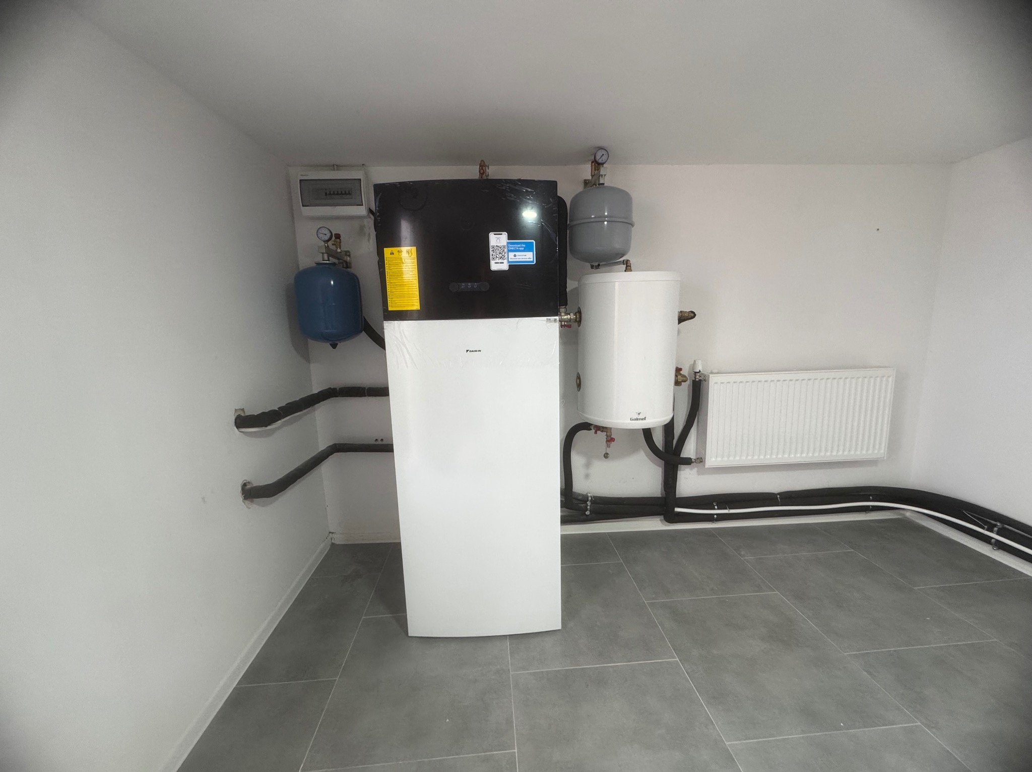 Nowoczesna instalacja z pompą ciepła Daikin, zbiornikiem buforowym i grzejnikiem panelowym w pomieszczeniu technicznym w Ligocie Małej, z widocznymi czarnymi rurami instalacyjnymi.