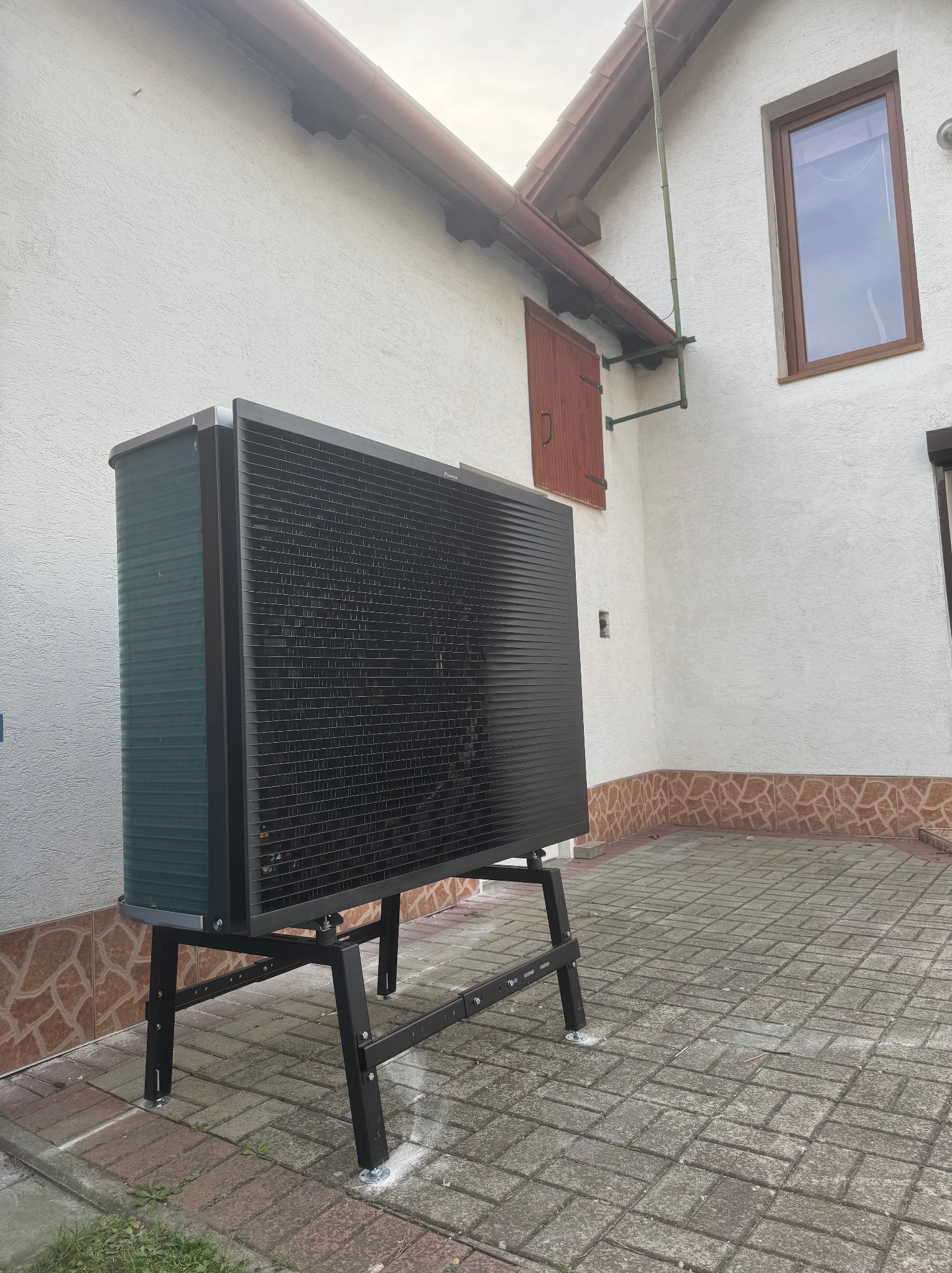 Czarna pompa ciepła powietrze-woda na stalowym stojaku, zainstalowana przy białej ścianie domu z brązowymi elementami wykończeniowymi, widoczna kostka brukowa.