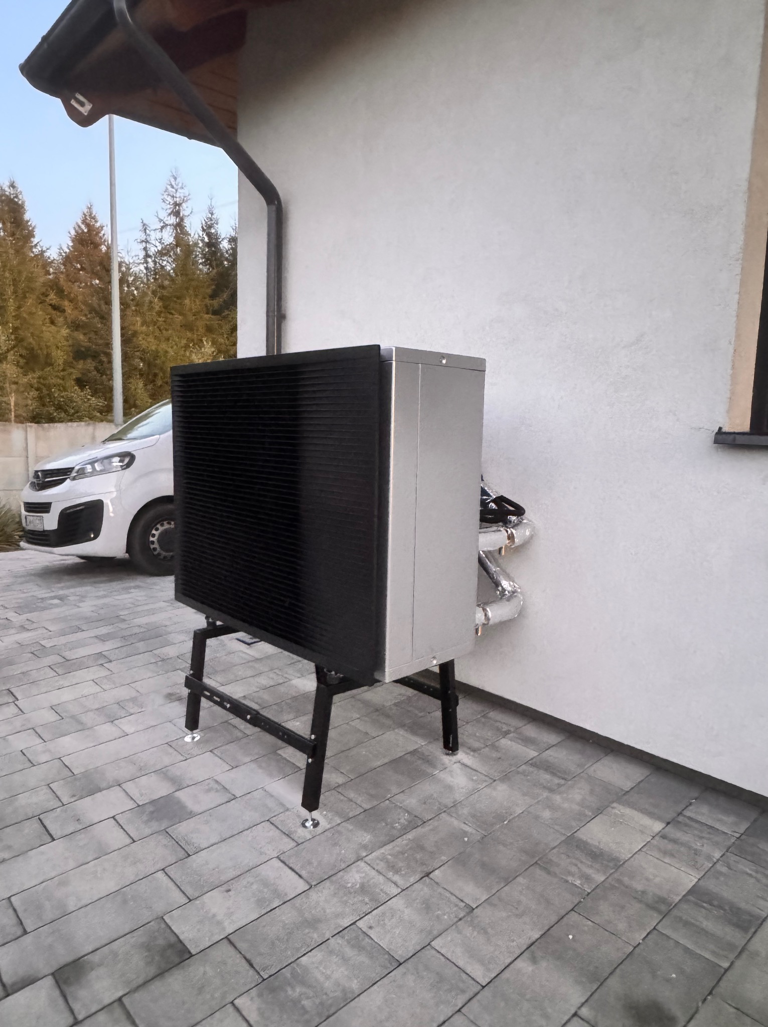 Zewnętrzna jednostka pompy ciepła powietrze-woda, zainstalowana przy ścianie budynku z widocznymi rurami w izolacji, na tle szarej kostki brukowej i białego samochodu.