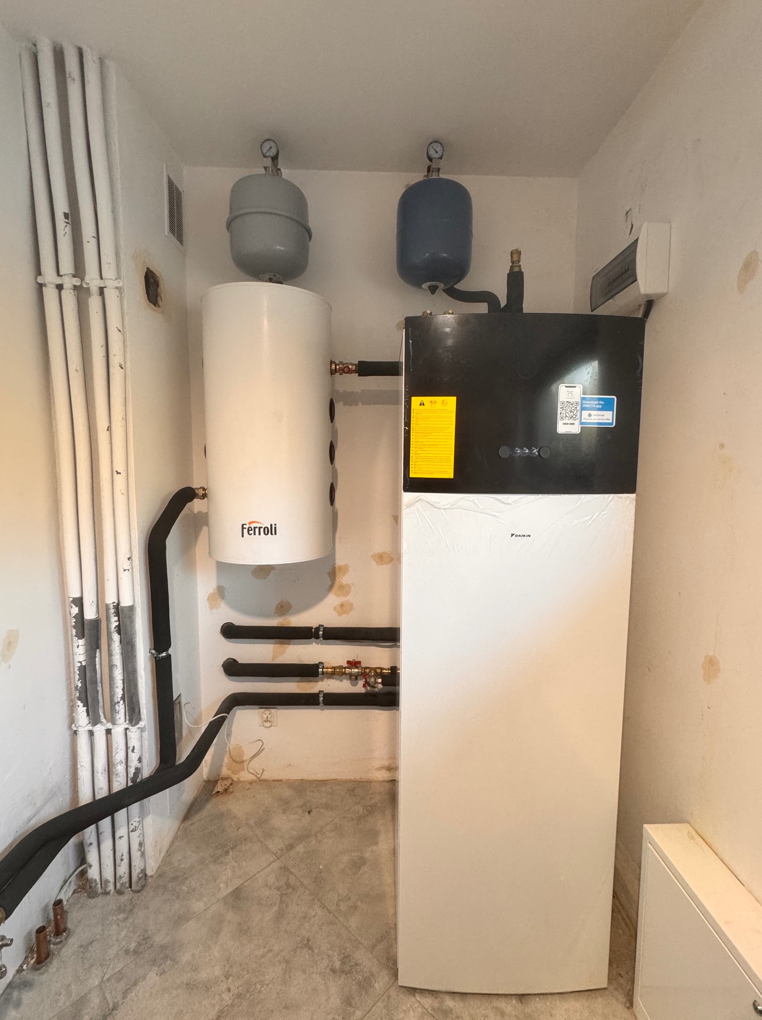 Nowoczesna instalacja grzewcza z zbiornikiem Ferroli i urządzeniem Daikin, rury w izolacji termicznej, manometry na zbiornikach, ściany z widocznymi śladami po instalacji.