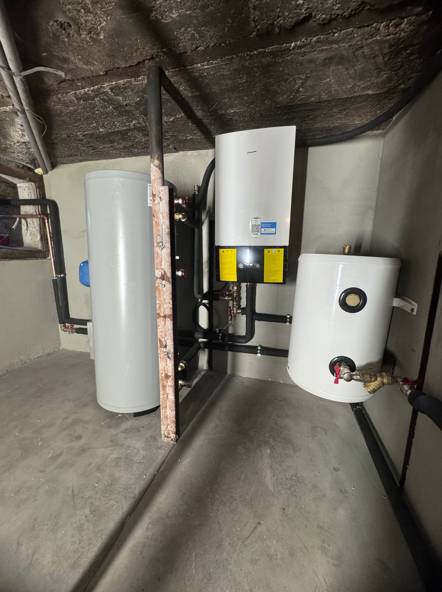Instalacja z białym zbiornikiem ciepłej wody, szarym zasobnikiem i pompą ciepła Daikin na tle surowego betonowego sufitu w pomieszczeniu technicznym.