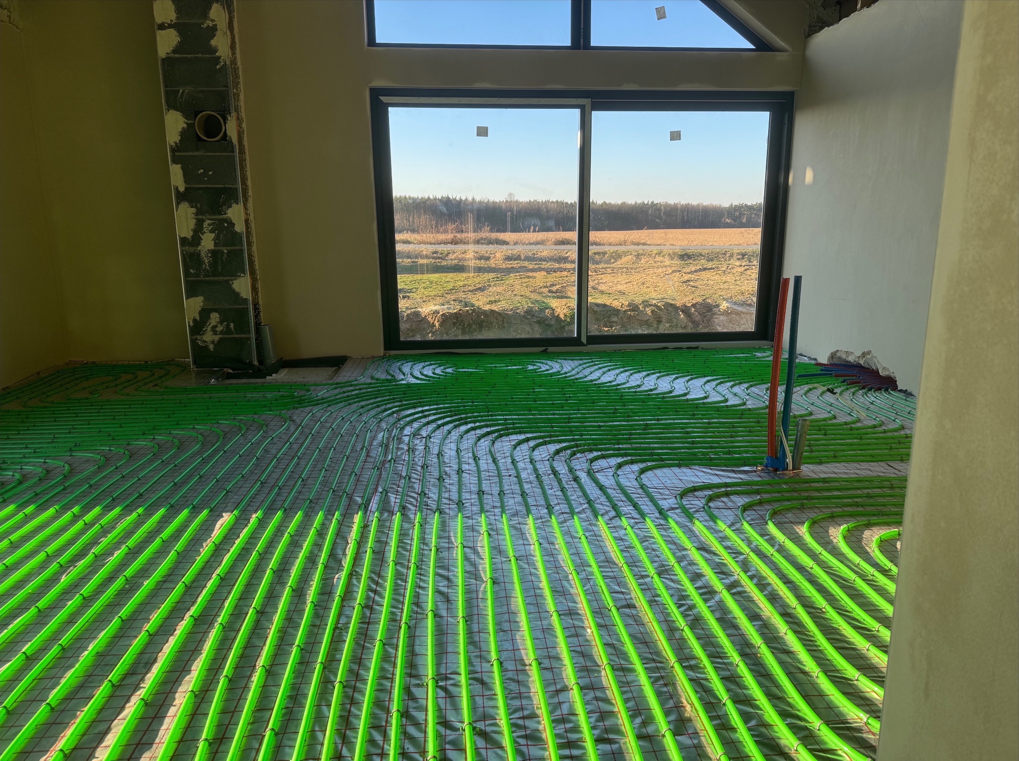 Wnętrze w stanie surowym z widoczną instalacją ogrzewania podłogowego z rur w kolorze jaskrawej zieleni, ułożonych na siatce w nowo budowanym domu z dużym oknem na krajobraz.