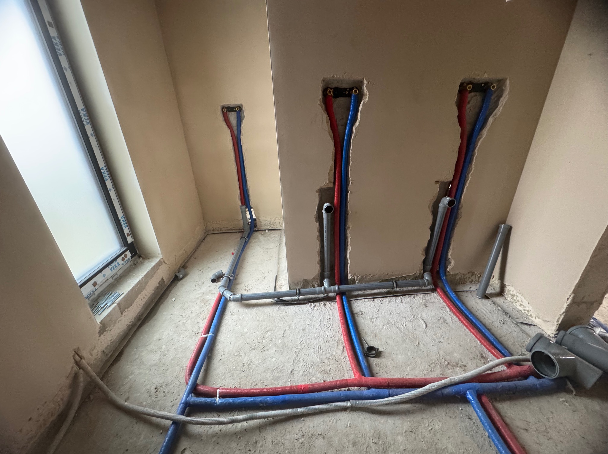 Surowe wnętrze w trakcie prac hydraulicznych: widoczne czerwone i niebieskie rury prowadzone w bruzdach ściennych oraz wzdłuż podłogi, szare rury PCV, okno w tle, ślady zaprawy na podłodze.