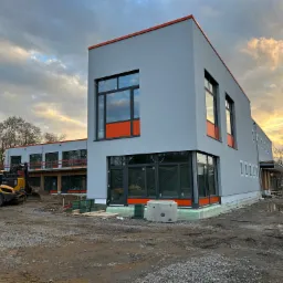 Nowoczesny budynek w trakcie budowy, szara elewacja z pomarańczowymi akcentami, duże okna, ziemia i sprzęt budowlany na pierwszym planie, zachodzące słońce w tle.