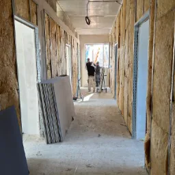 Korytarz w trakcie prac wykończeniowych: ściany z izolacją wełną mineralną, metalowe profile, stos płyt gipsowo-kartonowych, dw&oacute;ch pracownik&oacute;w montujących okno w oddali.