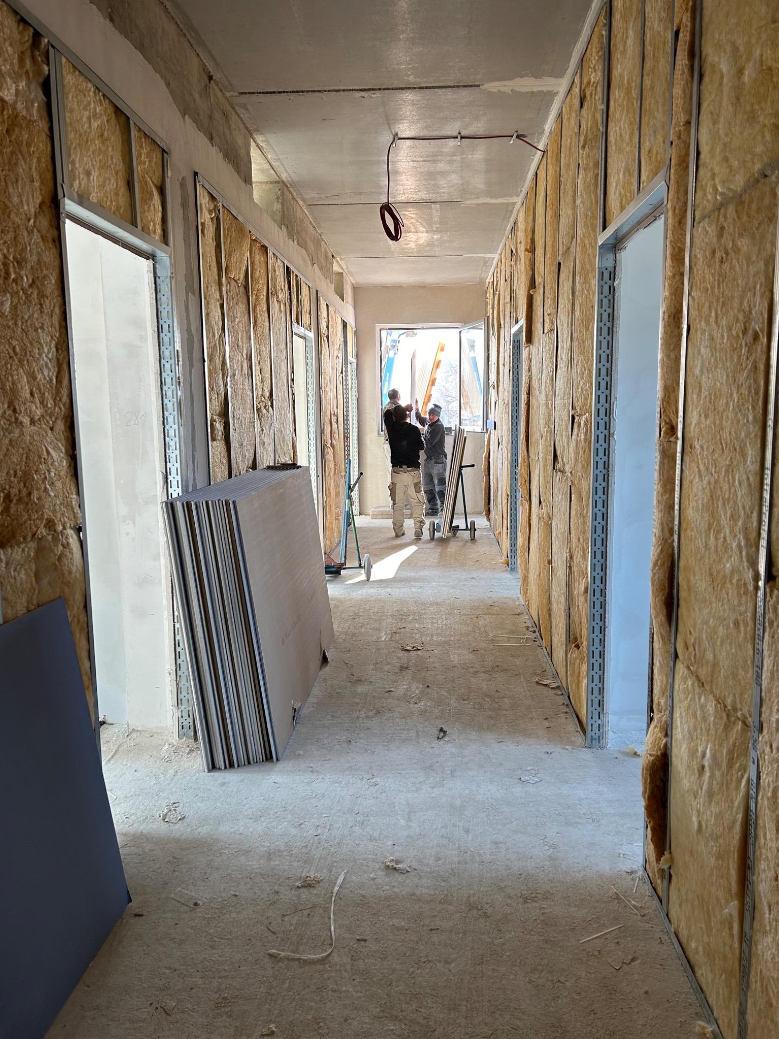 Korytarz w trakcie prac wykończeniowych: ściany z izolacją wełną mineralną, metalowe profile, stos płyt gipsowo-kartonowych, dwóch pracowników montujących okno w oddali.