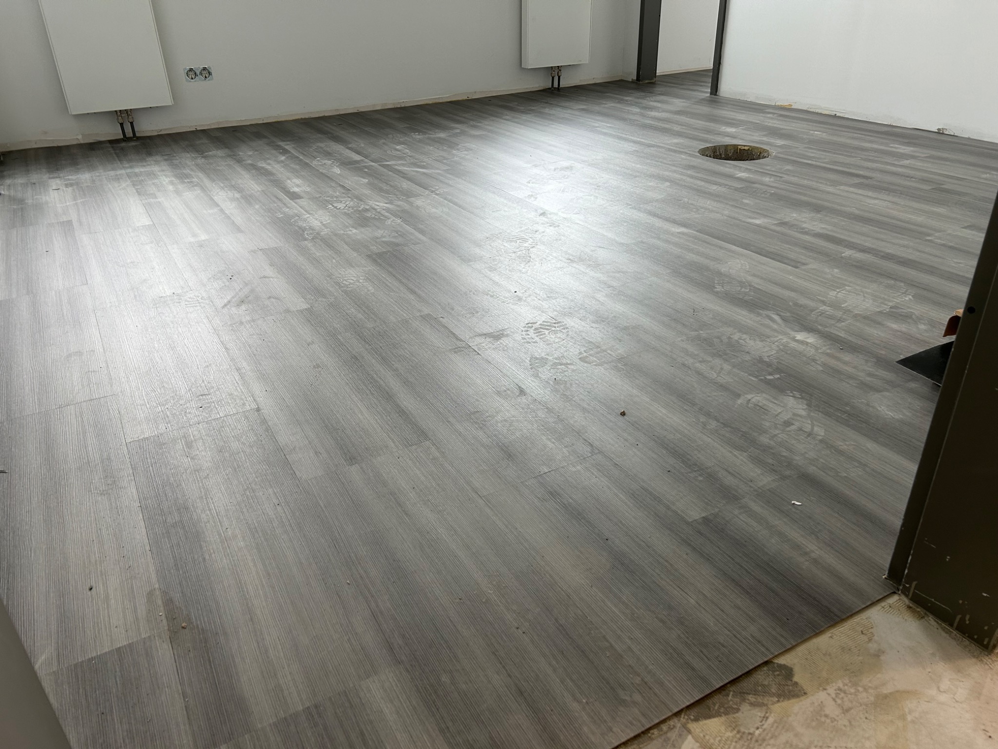 Świeżo położona, szara wykładzina podłogowa imitująca drewno w jasnym pomieszczeniu, widoczne ślady butów i otwór w podłodze.
