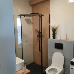 Nowoczesna łazienka z kabiną prysznicową typu walk-in z czarną ramą, toaletą podwieszaną, umywalką nablatową na drewnianym blacie i płytkami imitującymi drewno ułożonymi w jodełkę na ścianie...