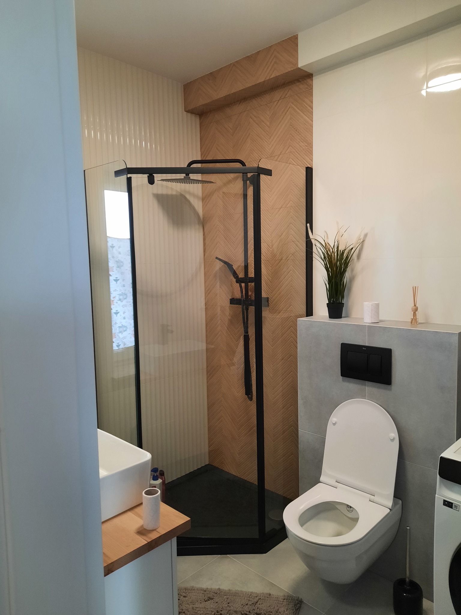 Nowoczesna łazienka z kabiną prysznicową typu walk-in z czarną ramą, toaletą podwieszaną, umywalką nablatową na drewnianym blacie i płytkami imitującymi drewno ułożonymi w jodełkę na ścianie...