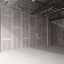 QuexTerm - Wnętrze w stanie surowym: ściany wyłożone płytami gipsowo-kartonowymi, widoczne łączenia płyt, instalacje pod sufitem, betonowa podłoga.