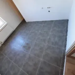 Szare płytki podłogowe ułożone w nowej łazience, widoczne przyłącza wodne na ścianie, grzejnik pod oknem.