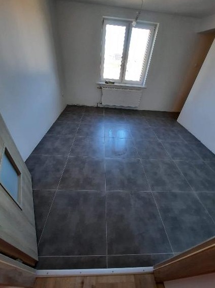 Pokój po remoncie z nową, ciemnoszarą podłogą z płytek, widoczne białe ściany, okno z grzejnikiem i fragment drzwi.