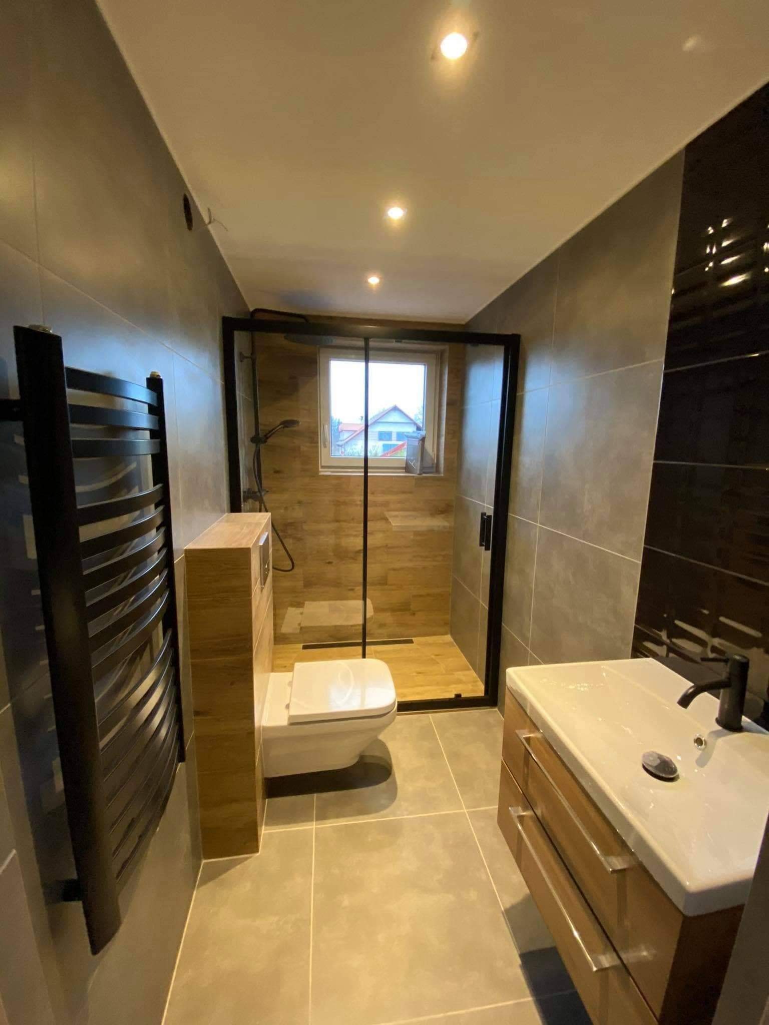 Nowoczesna łazienka z szarymi płytkami, kabiną prysznicową z czarnymi ramami, drewnopodobnymi akcentami na ścianach w kabinie i obudowie toalety, oraz grzejnikiem drabinkowym w kolorze czarnym.