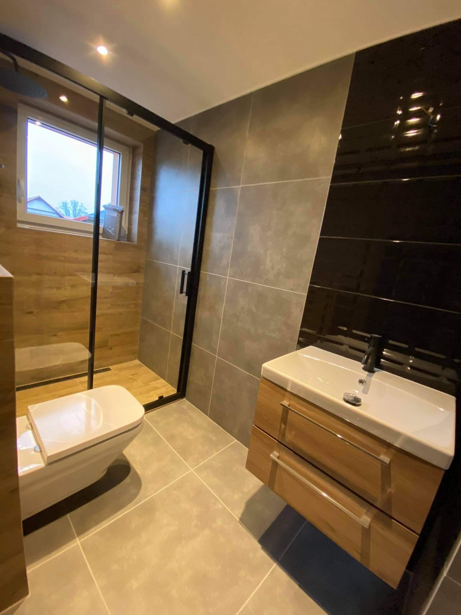 Nowoczesna łazienka z kabiną prysznicową typu walk-in, wiszącą toaletą i szafką pod umywalkę z drewnopodobnym fornirem oraz czarną armaturą.