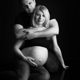 Paweł Otto – Fotograf Łódź | Studio Fotograficzne - Czarno-biała fotografia pary oczekującej dziecka. Kobieta w jeansach i topie, siedzi, trzymając dłoń na brzuchu. Mężczyzna obejmuje ją od tyłu. Ciemne tło.