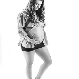 Paweł Otto – Fotograf Łódź | Studio Fotograficzne - Czarno-białe, studyjne zdjęcie kobiety w ciąży, ubrana w rozpiętą koszulę i szorty, delikatnie dotyka brzucha, z głową pochyloną w dół, na jasnym tle.