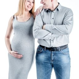 Paweł Otto – Fotograf Łódź | Studio Fotograficzne - Uśmiechnięta, przyszła mama w dopasowanej sukience i jej partner, pozują na białym tle, tworząc ciepłą, rodzinną atmosferę. Kobieta trzyma dłoń na brzuchu.