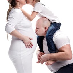 Paweł Otto – Fotograf Łódź | Studio Fotograficzne - Czułe ujęcie: ciężarna mama w wianku całowana przez synka, tata całuje brzuszek. Białe tło, jasne ubrania, rodzinna miłość. Sesja z bliska, skupienie na emocjach.
