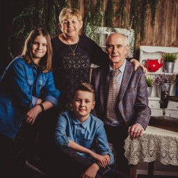 Paweł Otto – Fotograf Łódź | Studio Fotograficzne - Rodzinne zdjęcie portretowe w studio z drewnianym tłem, gdzie w tle widać regał z dekoracjami, a na pierwszym planie stoi stary stół z maszyną do szycia.