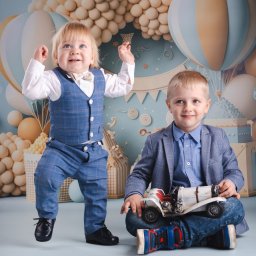 Paweł Otto – Fotograf Łódź | Studio Fotograficzne - Dwóch chłopców w eleganckich strojach pozuje na tle dekoracji z balonów i prezentów. Jeden stoi, drugi siedzi z zabawką-autkiem. Uśmiechnięte twarze, jasne tło.