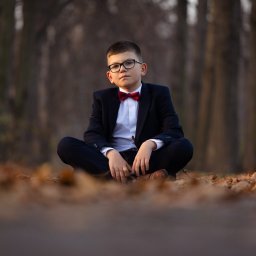 Paweł Otto – Fotograf Łódź | Studio Fotograficzne - Chłopiec w garniturze i muszce, siedzący po turecku na ziemi pokrytej liśćmi, na tle drzew. Portret w jesiennej scenerii.