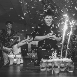 Paweł Otto – Fotograf Łódź | Studio Fotograficzne - Czarno-białe zdjęcie z urodzinowego przyjęcia: młody mężczyzna z tortem i fontannami iskier, konfetti w powietrzu, goście w tle, uśmiech, radosna atmosfera.