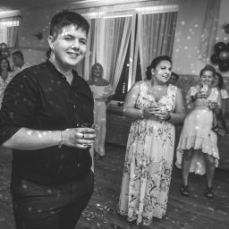 Paweł Otto – Fotograf Łódź | Studio Fotograficzne - Czarno-białe zdjęcie uśmiechniętego mężczyzny z drinkiem na tle świętujących osób podczas imprezy urodzinowej z dekoracjami i światłami dyskotekowymi.