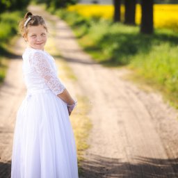 Paweł Otto – Fotograf Łódź | Studio Fotograficzne - Portret dziewczynki w białej sukience komunijnej na polnej drodze z kwitnącym rzepakiem w tle, delikatne światło, naturalna sceneria.
