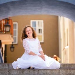 Paweł Otto – Fotograf Łódź | Studio Fotograficzne - Uśmiechnięta dziewczynka w białej sukience z koronką siedzi na schodach na tle architektury miejskiej. Jasne, naturalne światło.