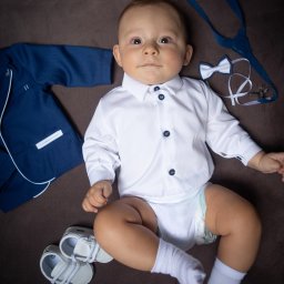 Paweł Otto – Fotograf Łódź | Studio Fotograficzne - Uroczy niemowlak w białej koszuli z granatowymi guzikami, leżący obok miniaturowego garnituru, muchy i bucików na brązowym tle.