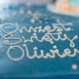 Paweł Otto – Fotograf Łódź | Studio Fotograficzne - Błękitny tort z białym napisem 'Chrzest Święty Oliwiera', zdobiony perłowymi koralikami i kwiatkami. Ujęcie z bliska, delikatne rozmycie tła.