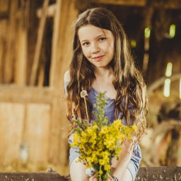 Paweł Otto – Fotograf Łódź | Studio Fotograficzne - Portret uśmiechniętej dziewczynki z bukietem polnych kwiatów w rustykalnej scenerii w Łodzi. Naturalne światło, drewniane tło i delikatny uśmiech.