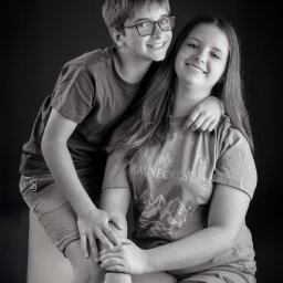 Paweł Otto – Fotograf Łódź | Studio Fotograficzne - Czarno-białe portretowe ujęcie uśmiechniętego rodzeństwa: nastolatki siedzącej z chłopcem opierającym się o jej ramię, na ciemnym tle.