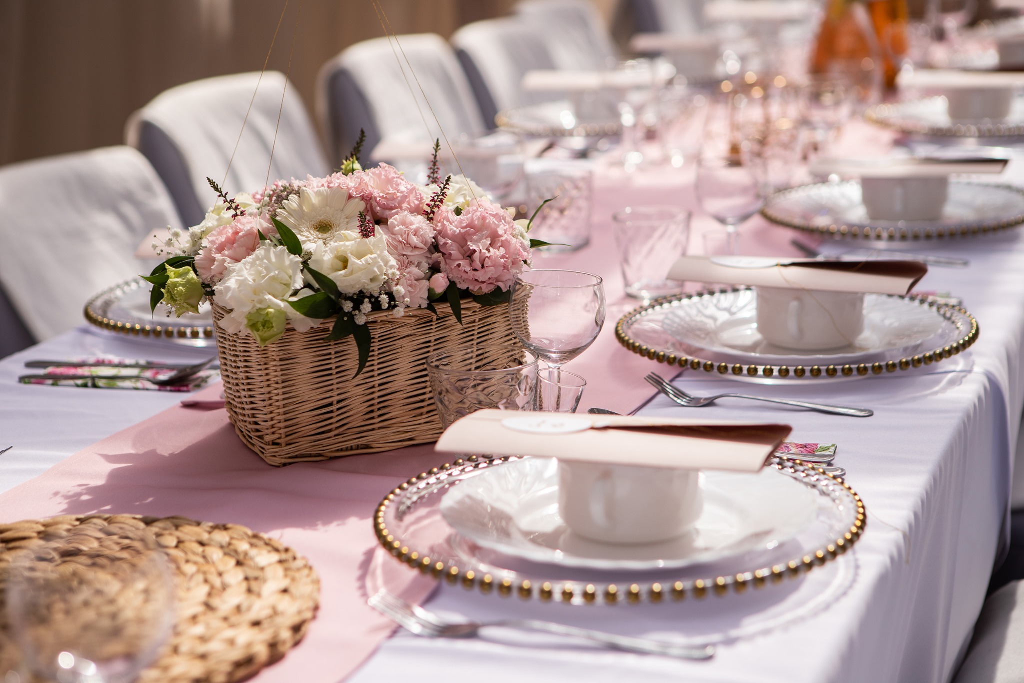 Uroczysty stół z kwiatami w koszyku, nakryty elegancką zastawą z dekoracyjnymi talerzami i delikatnym różowym bieżnikiem, przygotowany na przyjęcie.