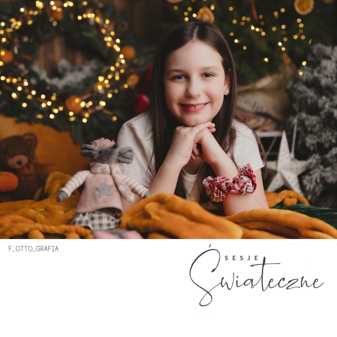 Uśmiechnięta dziewczynka z brązowymi włosami, oparta na rękach, w świątecznej scenerii z pluszowym misiem i dekoracjami.